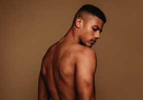 Close up of shirtless african american person looking down. Portrait of muscular standing on brown background.